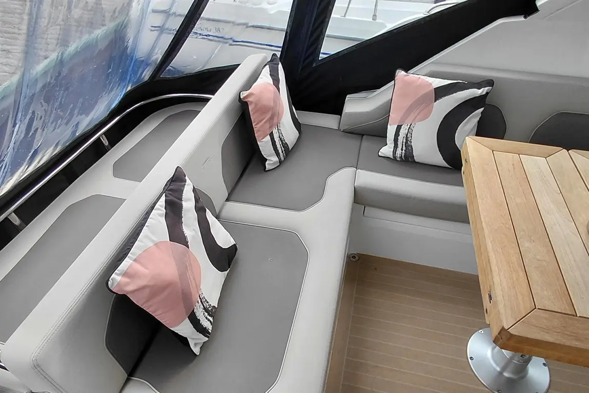 Stylish boat interior with gray seating, decorative pillows, and a wooden table, featuring a backdrop of marina views.